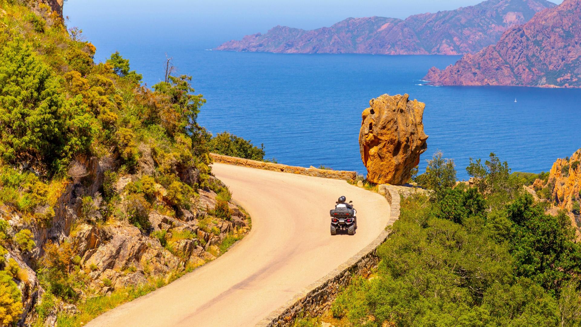 Strada panoramica costiera con moto, vegetazione rigogliosa e mare blu sullo sfondo.