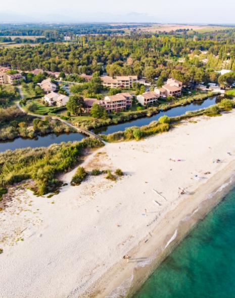 Spiaggia sabbiosa con mare turchese, vegetazione e case in lontananza.