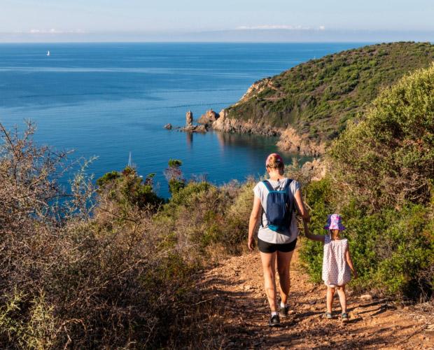 Madre e figlia camminano su un sentiero con vista mare e colline verdi.