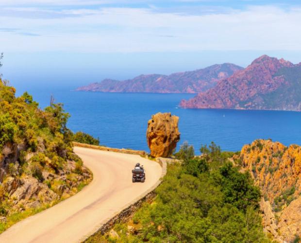 Strada panoramica con moto, vista mare e montagne rosse in lontananza.