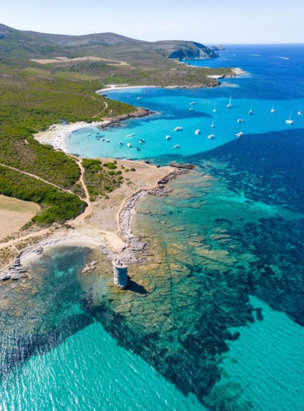 Spiaggia con torre antica e mare turchese, barche ancorate in baia.